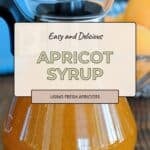 Glass pitcher filled with homemade apricot syrup on a wooden table, with fresh apricots in the background. This sweet apricot syrup is perfect for drizzling over desserts or mixing into summer drinks.