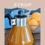 A glass syrup dispenser filled with apricot syrup sits on the table, with a bowl of fresh apricots in the background.
