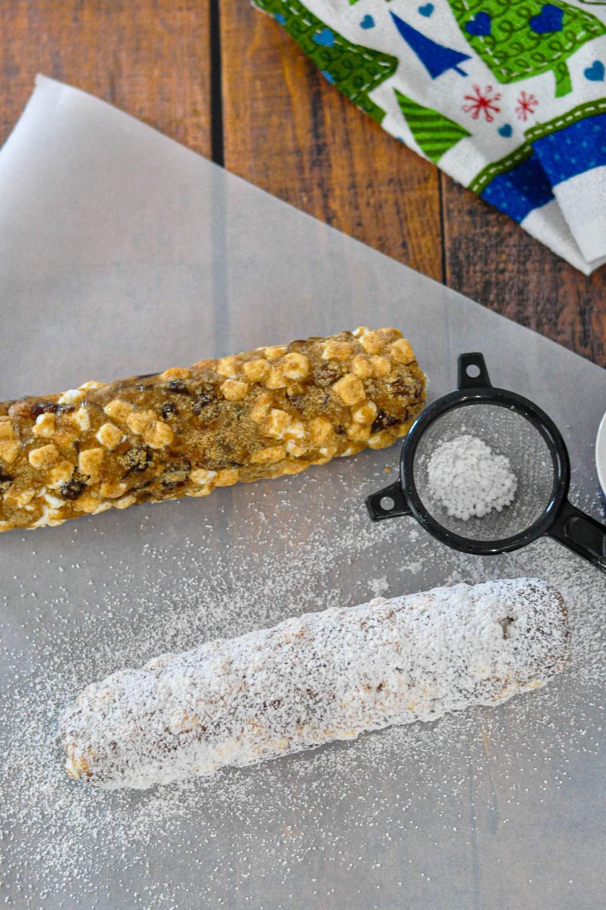 Two log-shaped desserts, one plain and one dusted with powdered sugar, rest on parchment with a sifter nearby-classic treats reminiscent of an old fashioned date roll, perfect for any holiday gathering.