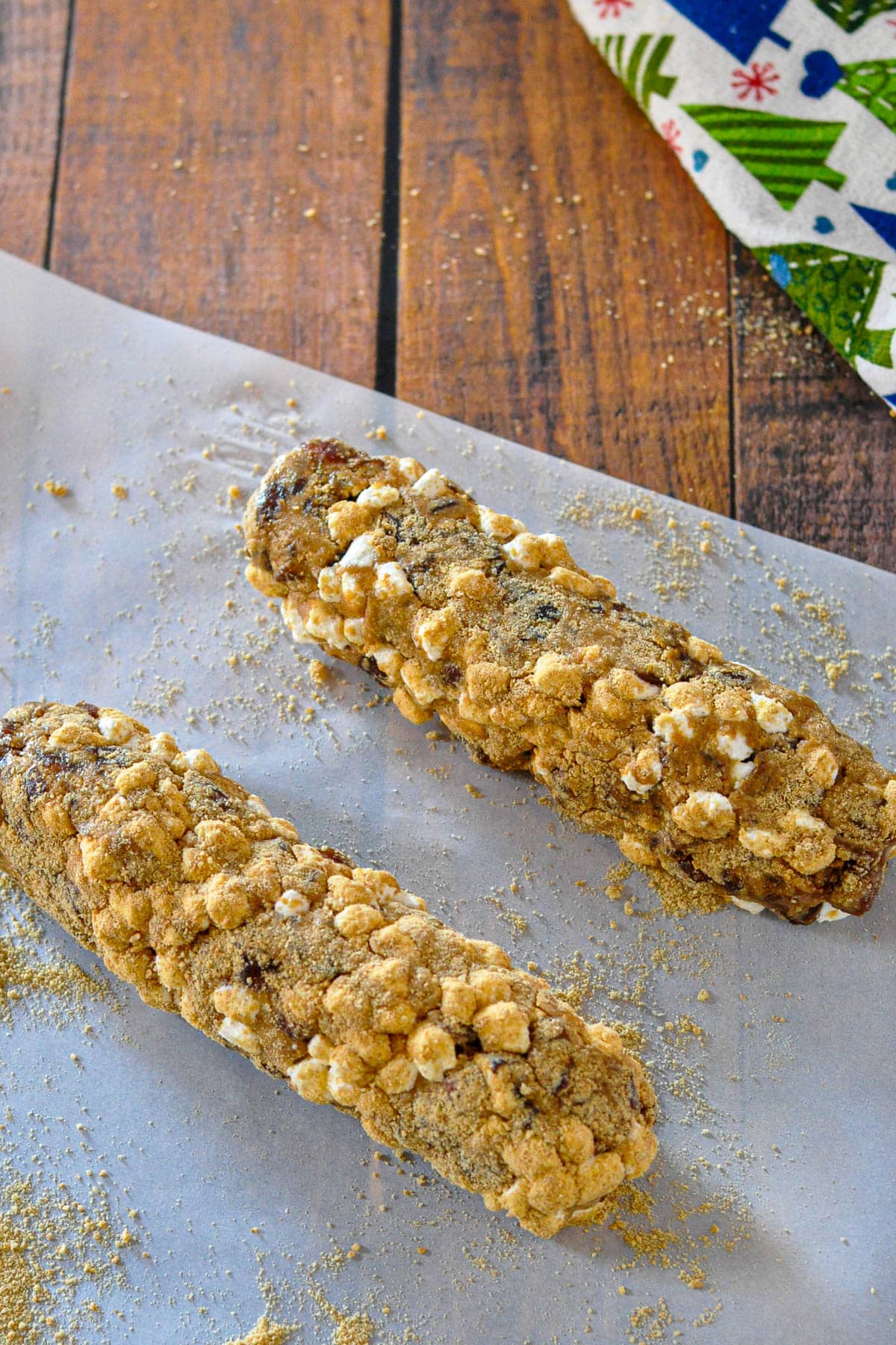 Two log-shaped Holiday Date Roll desserts topped with mini marshmallows and crumbs rest on parchment paper, set atop a wooden surface.