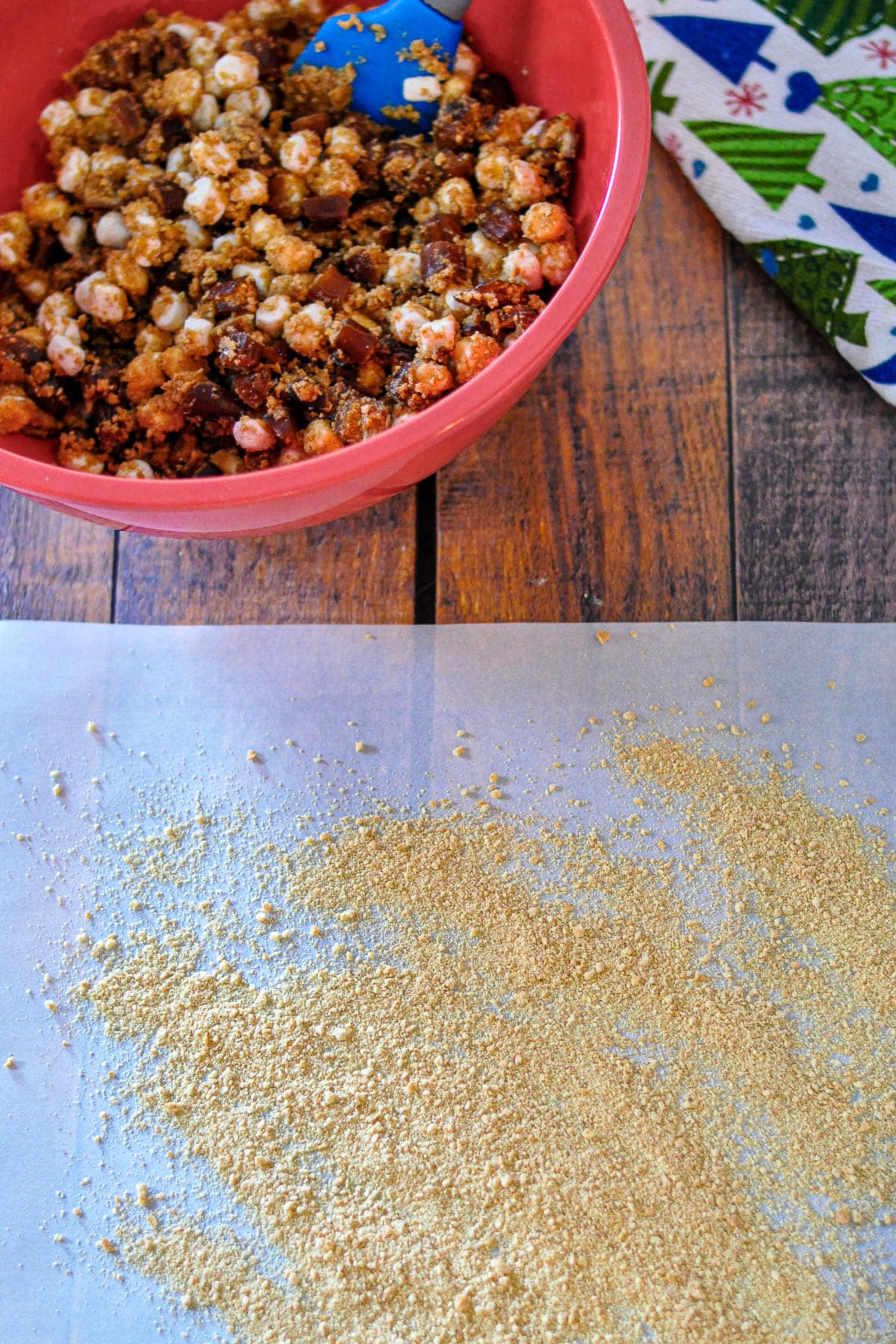 A red bowl of snack mix with marshmallows and a blue spatula sits beside a sheet of crushed graham crackers, ready for making a classic Holiday Date Roll.