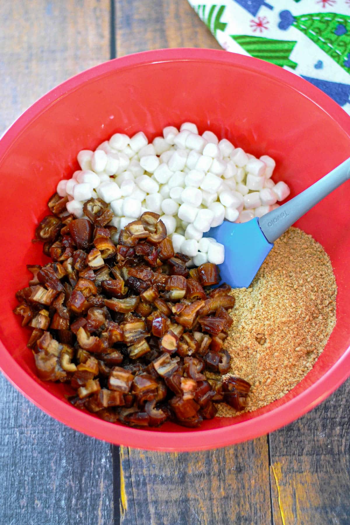 Red bowl with mini marshmallows, chopped dates, and graham cracker crumbs mixed with a blue spatula-the perfect start for an old fashioned date roll or classic Holiday Date Roll treat.