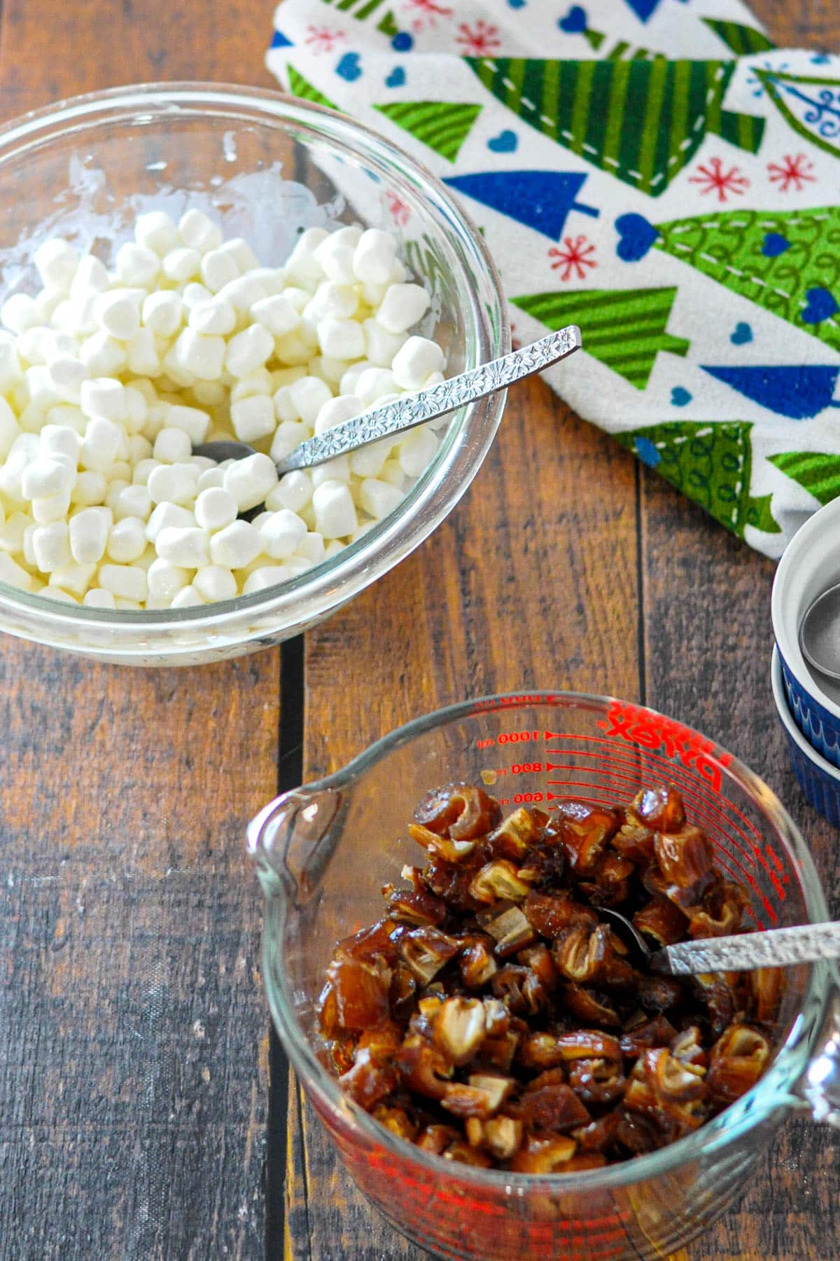 A bowl of mini marshmallows and a measuring cup of chopped dates sit on a wooden table with a festive napkin, ready to make an old fashioned date roll for the holidays.