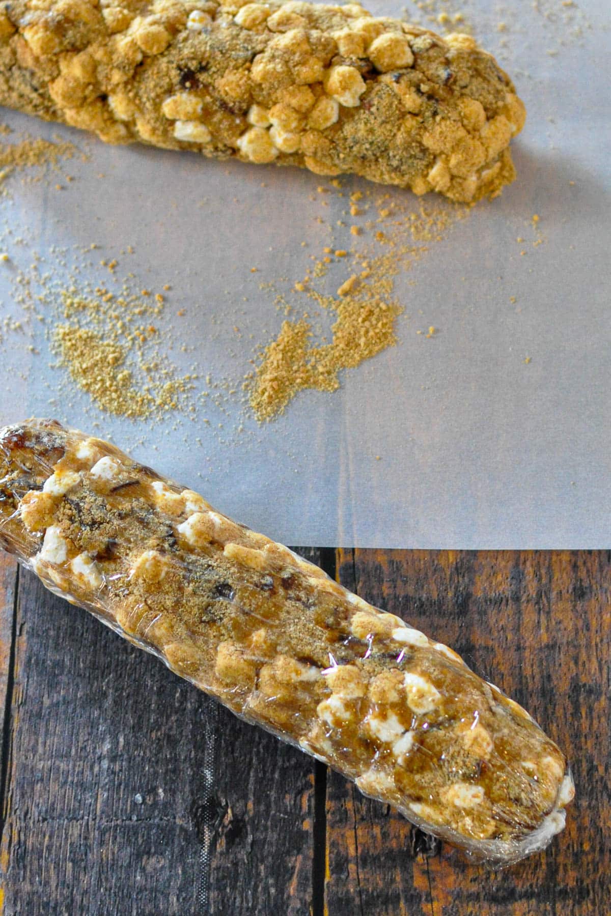 Two logs of Holiday Date Roll dessert, filled with marshmallows and crumbs-one resting on parchment paper, the other on a wooden surface-bring old fashioned date roll charm to your table.