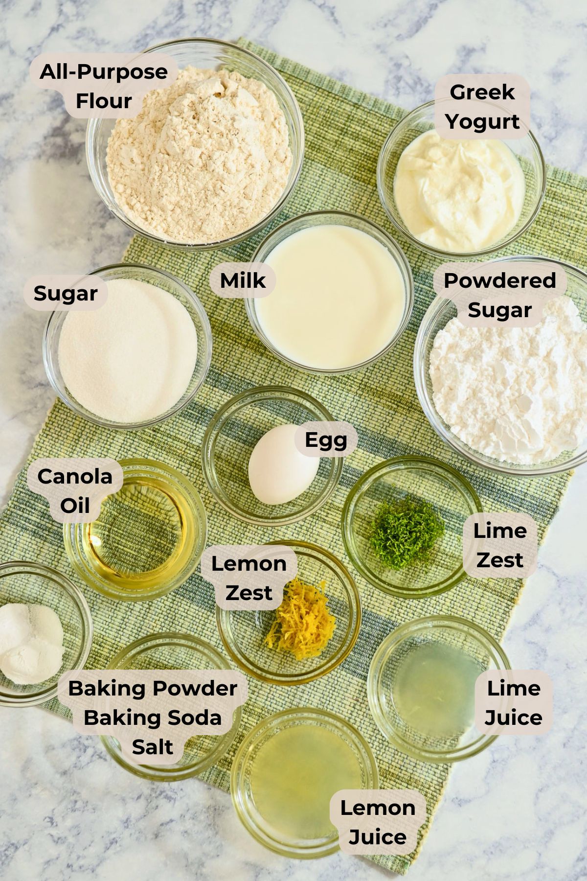 Ingredients for Lemon Lime Muffins arranged in bowls on a green mat, including flour, yogurt, milk, sugars, egg, oils, zests, juice, and leaveners.