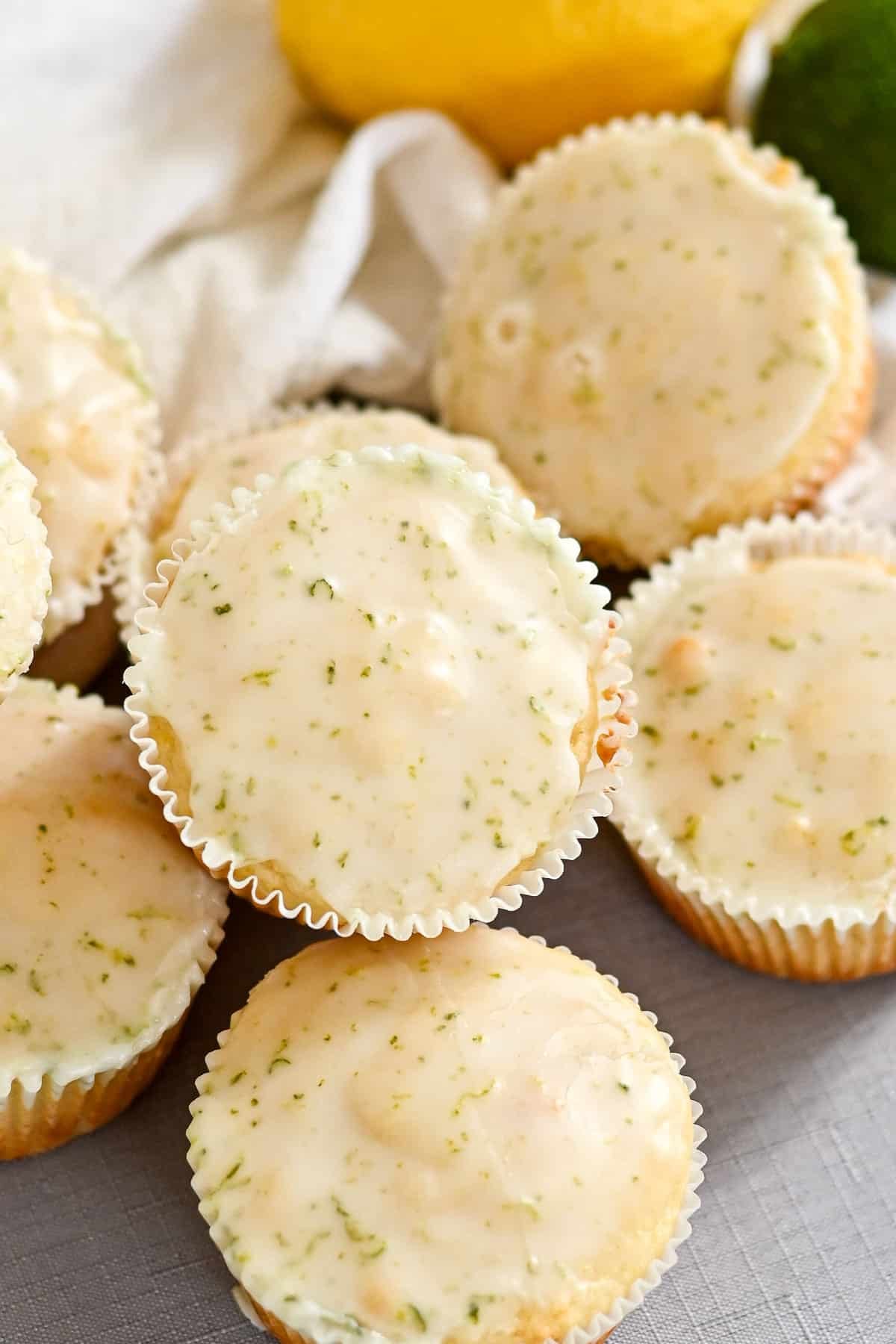 Lemon and lime glazed cupcakes, reminiscent of Lemon Lime Muffins, are arranged on a light surface with citrus fruits in the background.