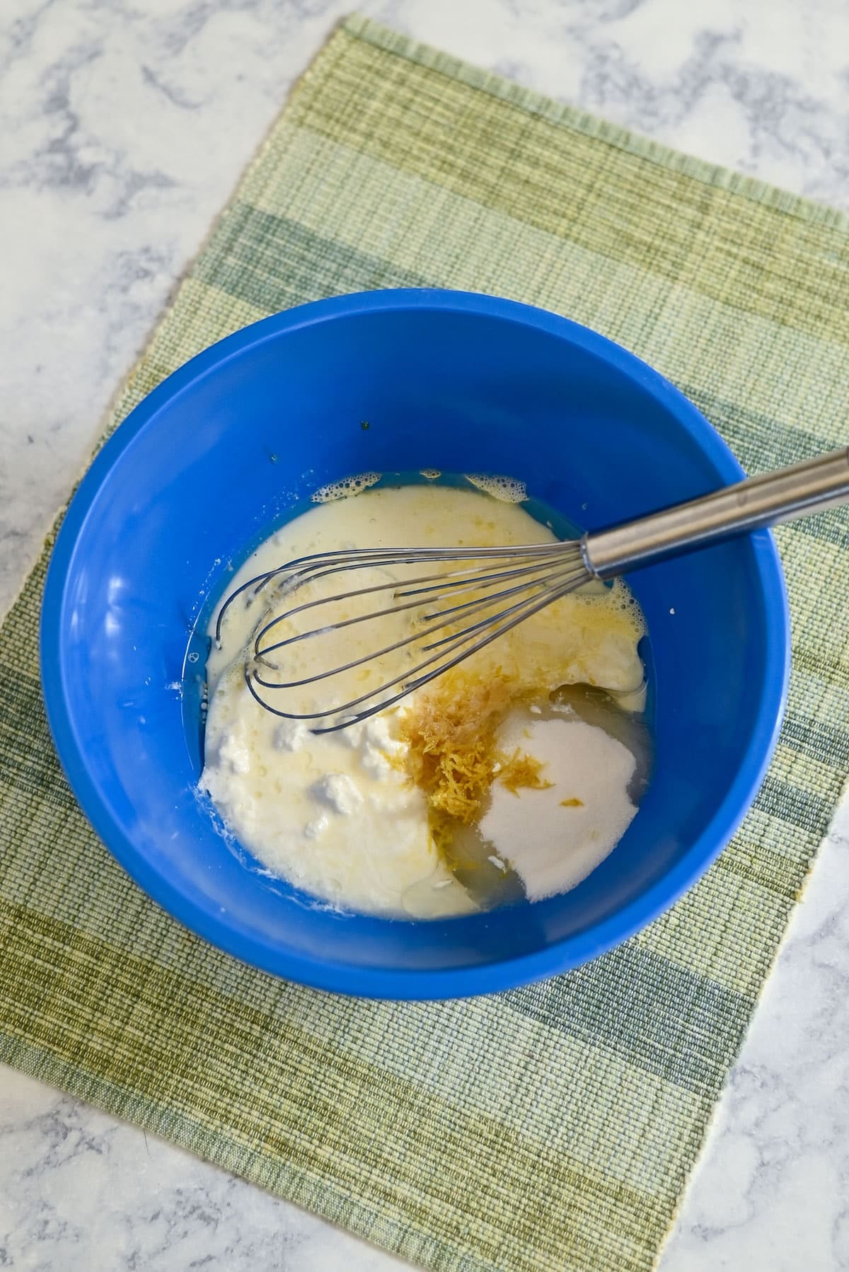 Blue bowl with whisk, containing sugar, lemon zest, and wet ingredients for Lemon Lime Muffins, on a green placemat and marble surface.