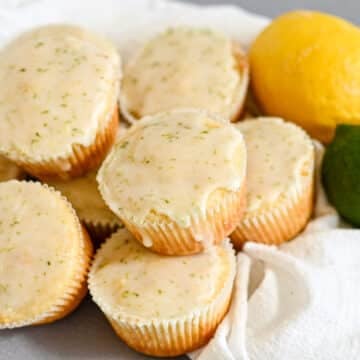 Lemon Lime Muffins glazed with citrusy zest are stacked on a white cloth, accompanied by fresh lemon and lime for a burst of flavor and color.