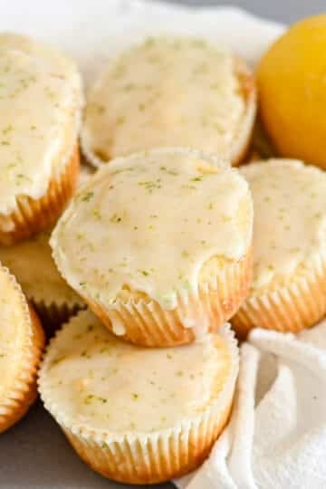 Lemon Lime Muffins glazed with citrusy zest are stacked on a white cloth, accompanied by fresh lemon and lime for a burst of flavor and color.