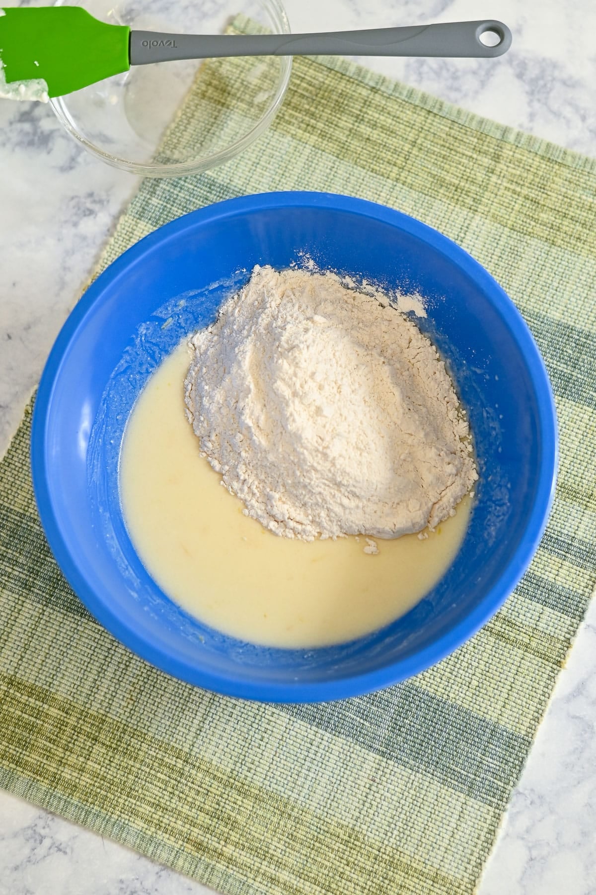 Blue bowl with flour and liquid mixture for Lemon Lime Muffins on a green plaid mat, with a spatula and glass bowl nearby.