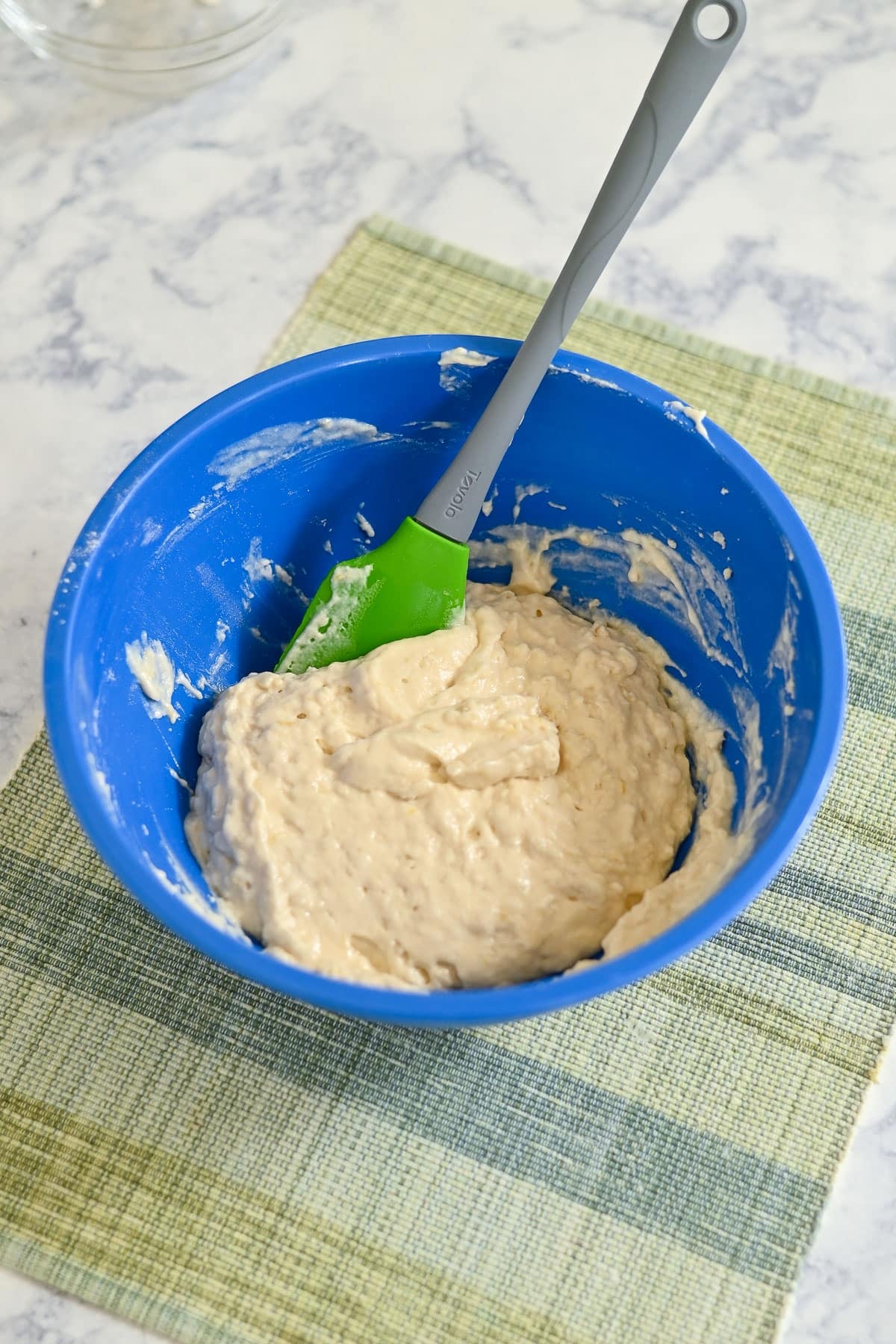 Blue bowl with thick Lemon Lime Muffins batter and a green spatula, sitting on a green plaid placemat on a marble surface.