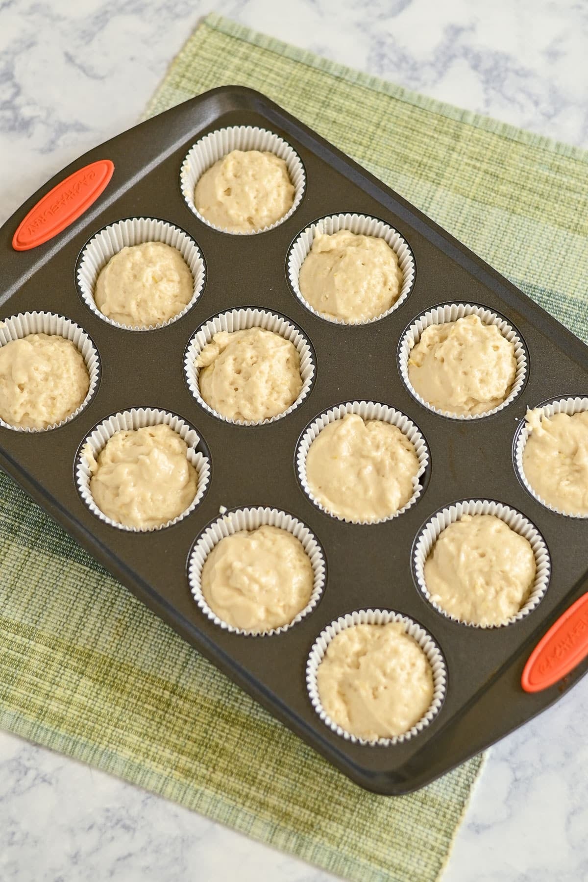 A muffin tin filled with cupcake liners and unbaked Lemon Lime Muffins batter, placed on a green checkered cloth.