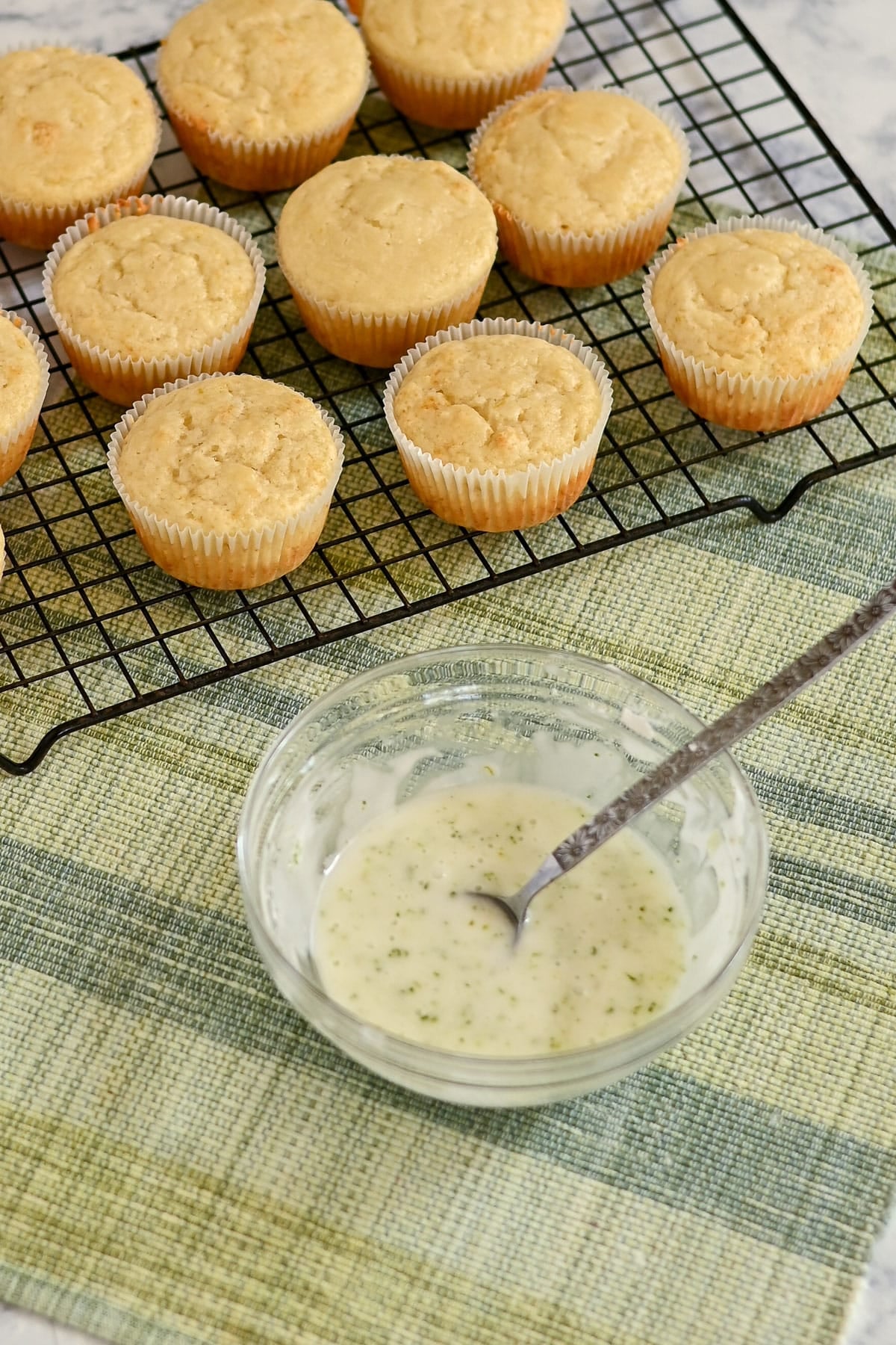 Lemon Lime Muffins cooling on a wire rack next to a bowl of icing with a spoon on a green placemat.