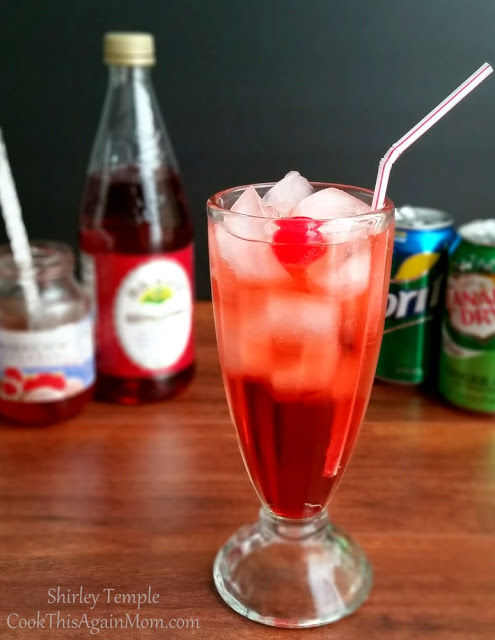 A tall glass filled with ice and a mixed shirley temple with the ingredients in the background