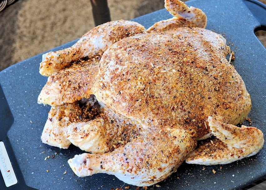 a raw whole chicken sitting on a black cutting board covered with seasoning