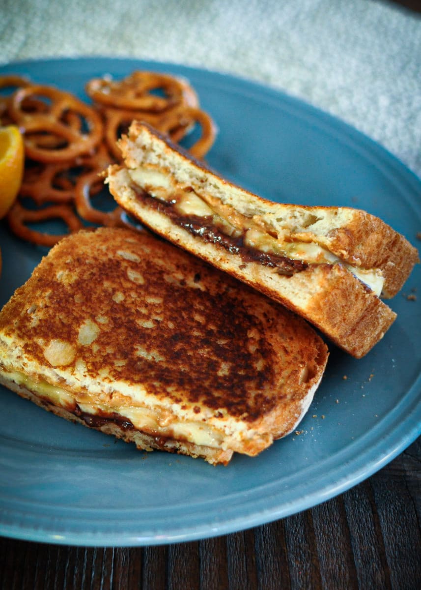 a grilled sandwich with peanut butter, Nutella, and banana sitting on a blue plate next to pretzels.