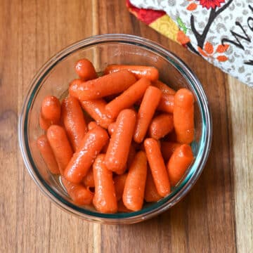 Glazed carrots in a clear glass bowl next to a kitchen towel.