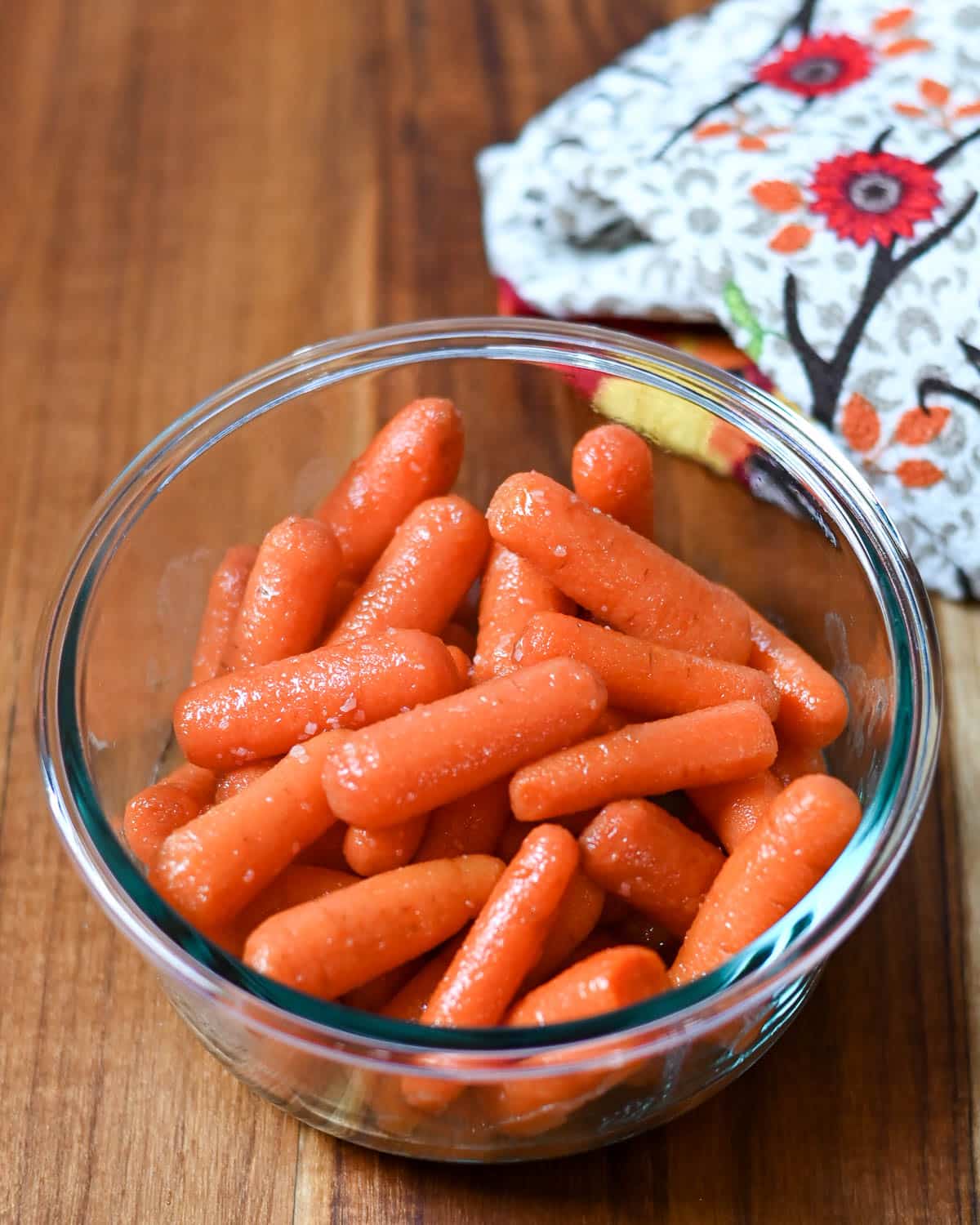 Glazed carrots in a clear glass bowl next to a kitchen towel.