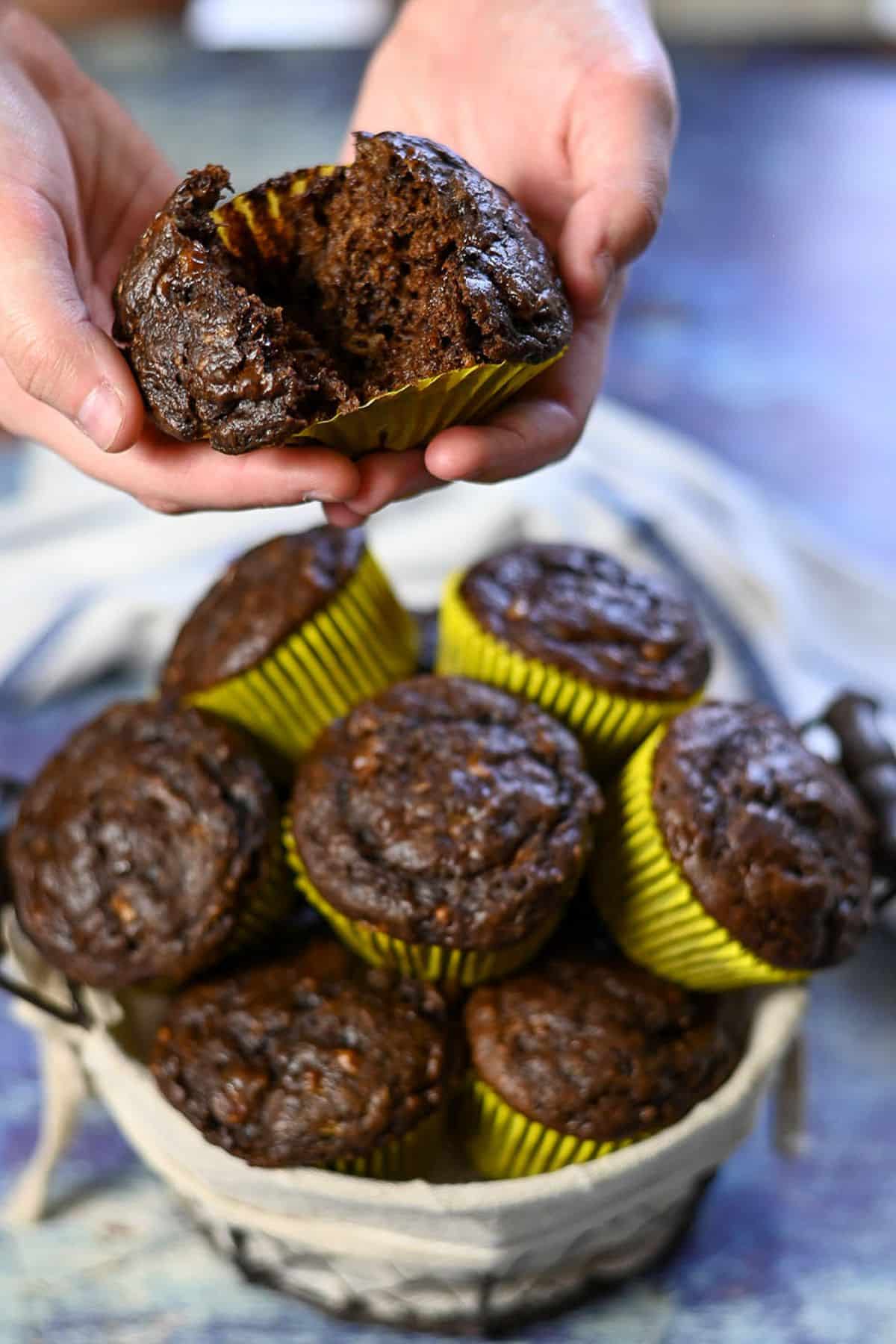 Hands holding a split chocolate banana muffin above a basket filled with chocolate muffins in yellow wrappers.
