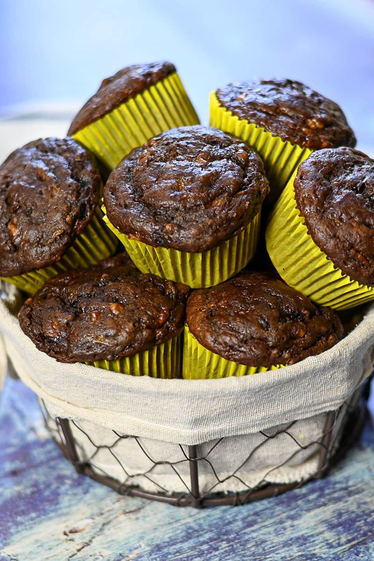 Chocolate Banana Muffins Basket filled with Chocolate Banana Muffins in yellow paper liners, set on a blue surface.