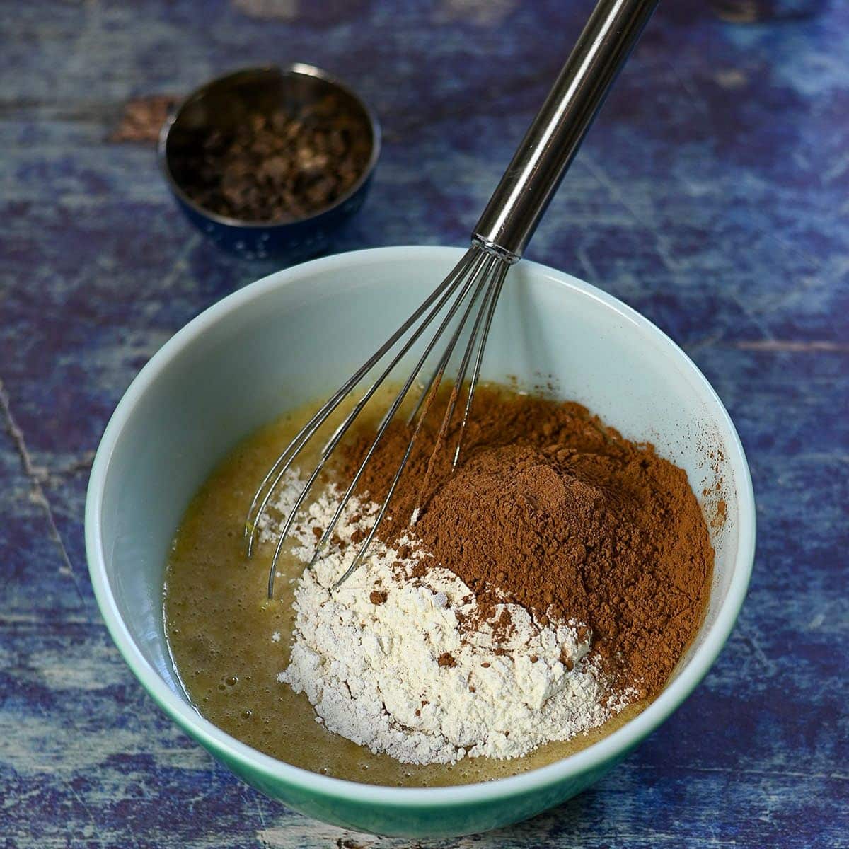 A bowl with flour, cocoa powder, and wet ingredients being mixed with a whisk for Chocolate Banana Muffins; a small bowl sits behind.