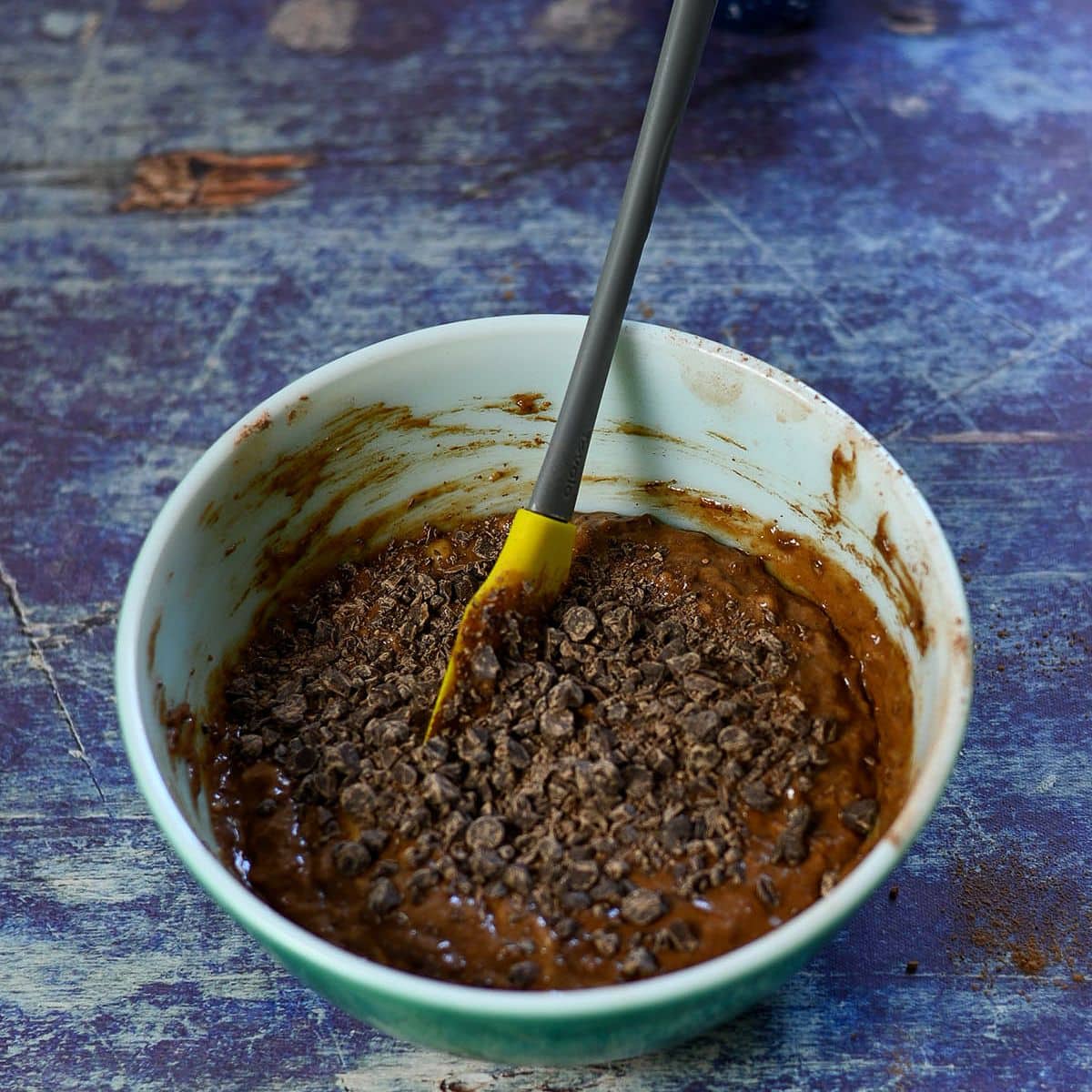 A mixing bowl with chocolate batter and chopped chocolate pieces for Chocolate Banana Muffins, stirred with a yellow spatula on a blue table.