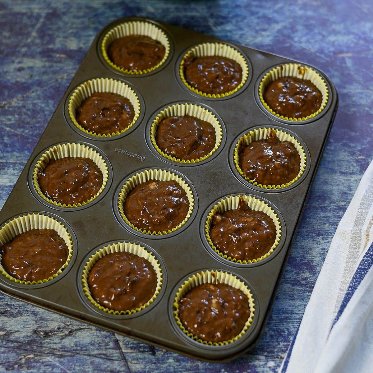 A muffin tin filled with chocolate banana muffins batter in yellow paper liners sits on a blue countertop.