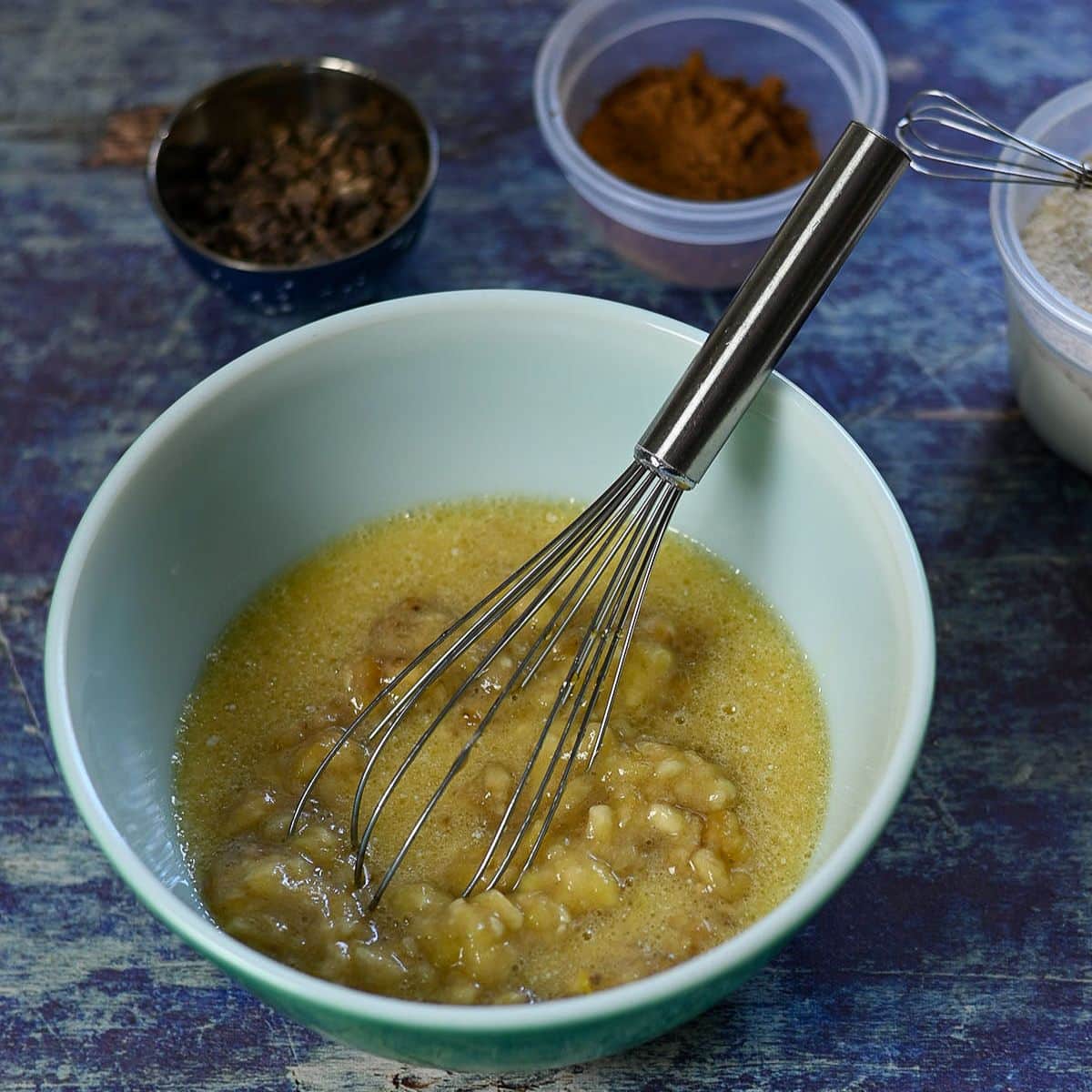 Mashed bananas and eggs being whisked in a bowl, with spices and chocolate in small containers nearby-perfect for preparing delicious Chocolate Banana Muffins.