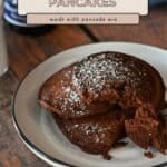A plate of three easy chocolate pancakes with cocoa powder, dusted with powdered sugar, sits on a wooden table.