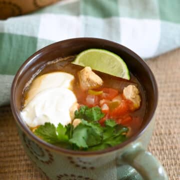A mug of Mexican chicken tortilla soup with sour cream, lime, cilantro, and diced vegetables sits on a woven mat.