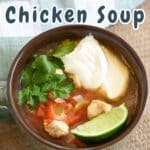 A steaming bowl of Mexican chicken soup sits on a rustic burlap surface, garnished with fresh lime, sprigs of cilantro, and a dollop of tangy sour cream.