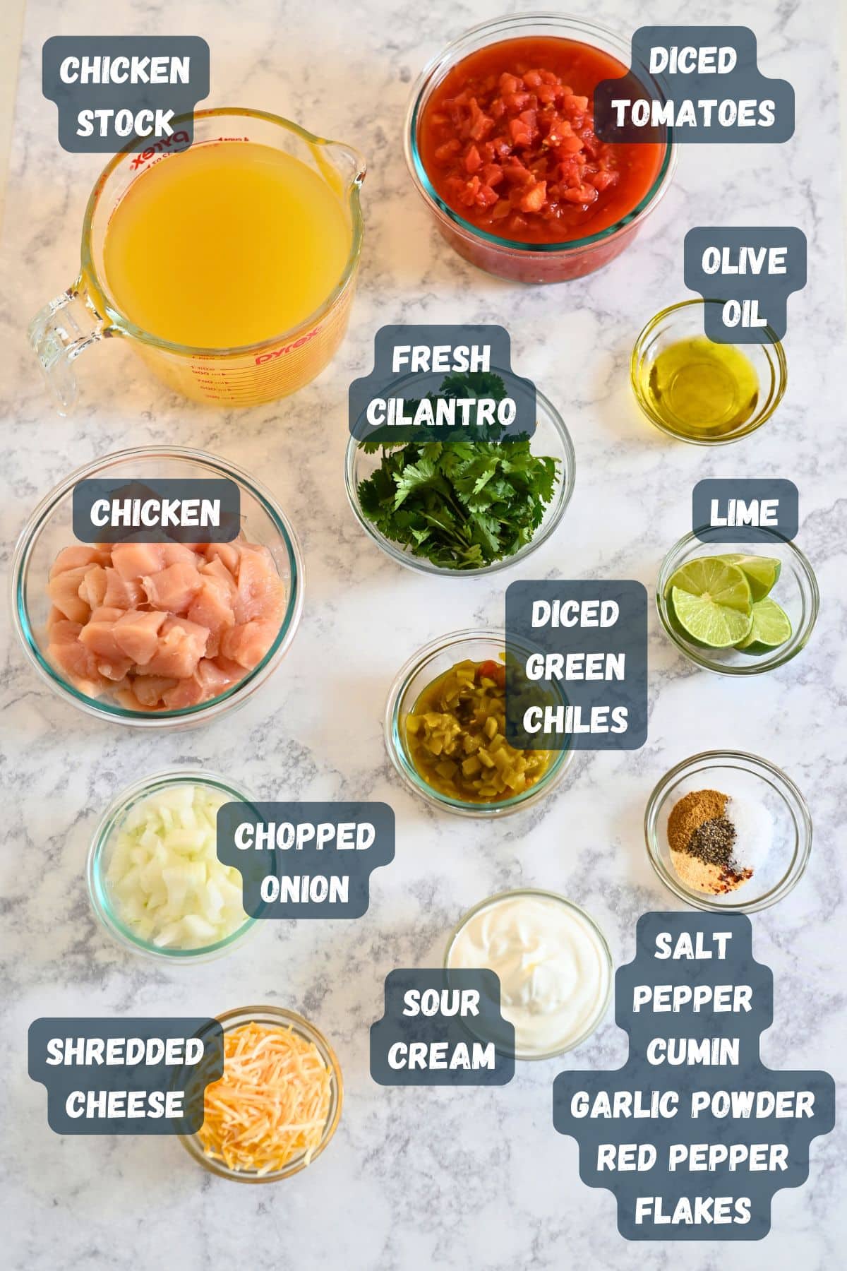 Ingredients for a delicious Mexican Chicken Soup are spread across a marble surface: chicken stock, tender chicken, fresh cilantro, ripe tomatoes, green chiles, cheese, and a variety of seasonings.