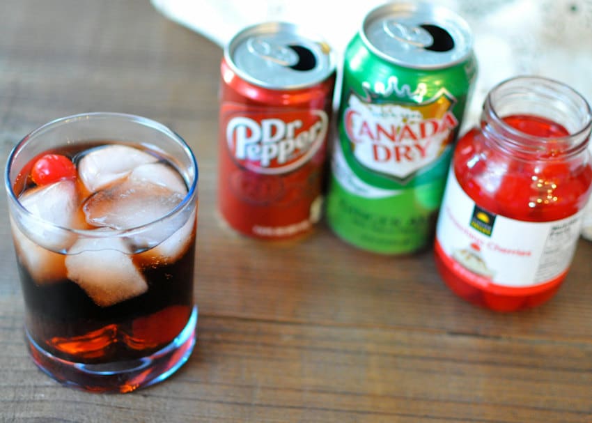 Dr. Pepper in a glass with ice and a cherry next to soda cans and a jar of cherries