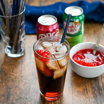 Soda with cherries next to a blue towel and a glass of straws.