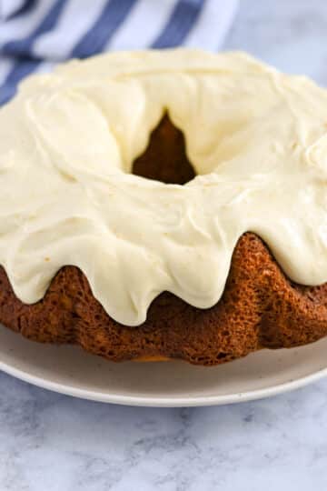 An orange cake with cream cheese frosting sits elegantly on a white plate, placed on a marble surface. A striped cloth is in the background.