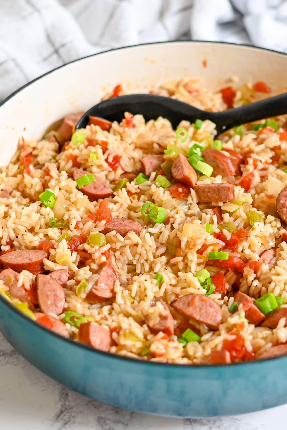 A pan of savory sausage jambalaya featuring Hillshire Farm smoked sausage, rice, tomatoes, and green onions with a black serving spoon resting inside.