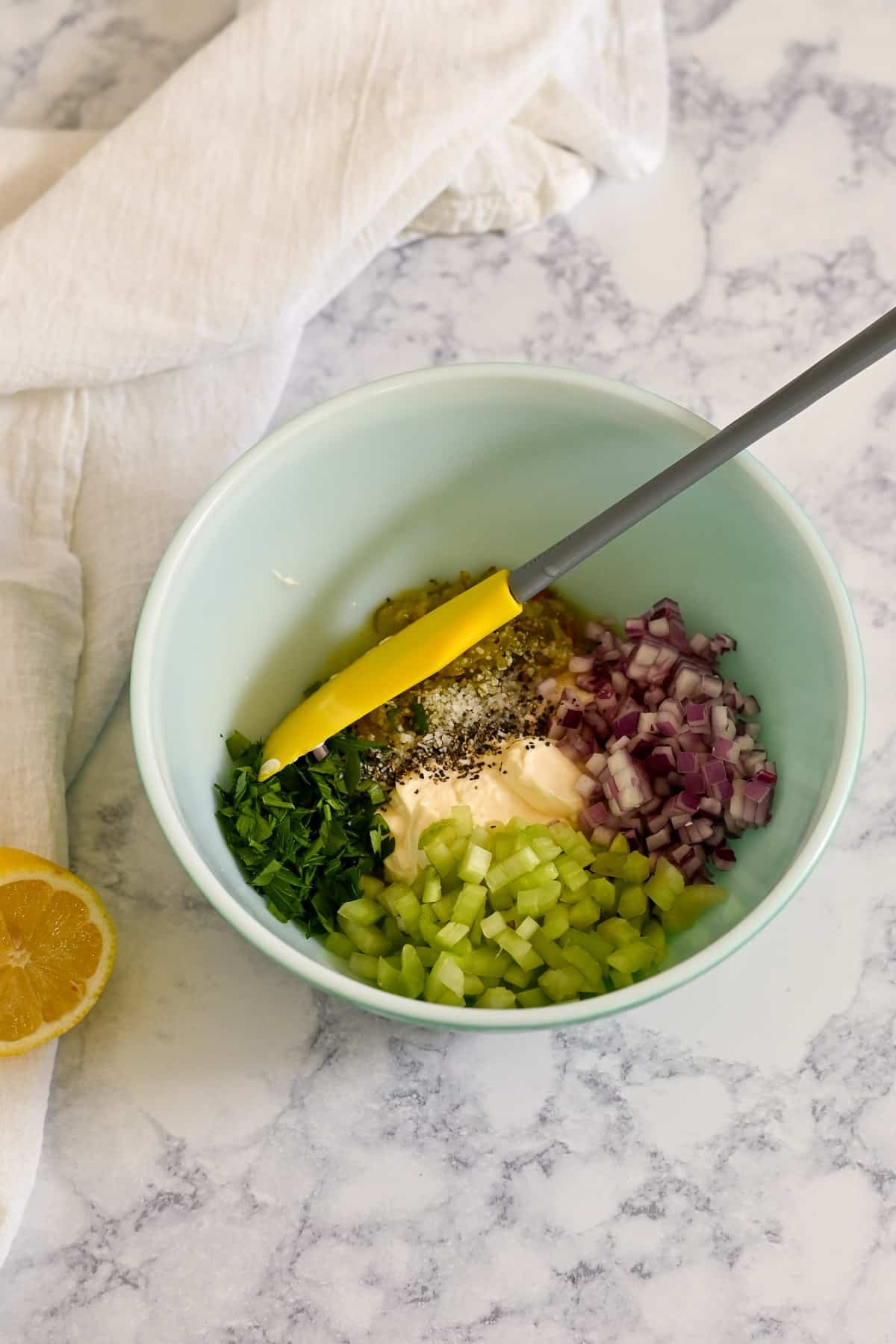 A mixing bowl with chopped celery, red onion, herbs, mayo, and a spatula on a marble counter-perfect for preparing a flavorful Rotisserie Chicken Macaroni Salad.
