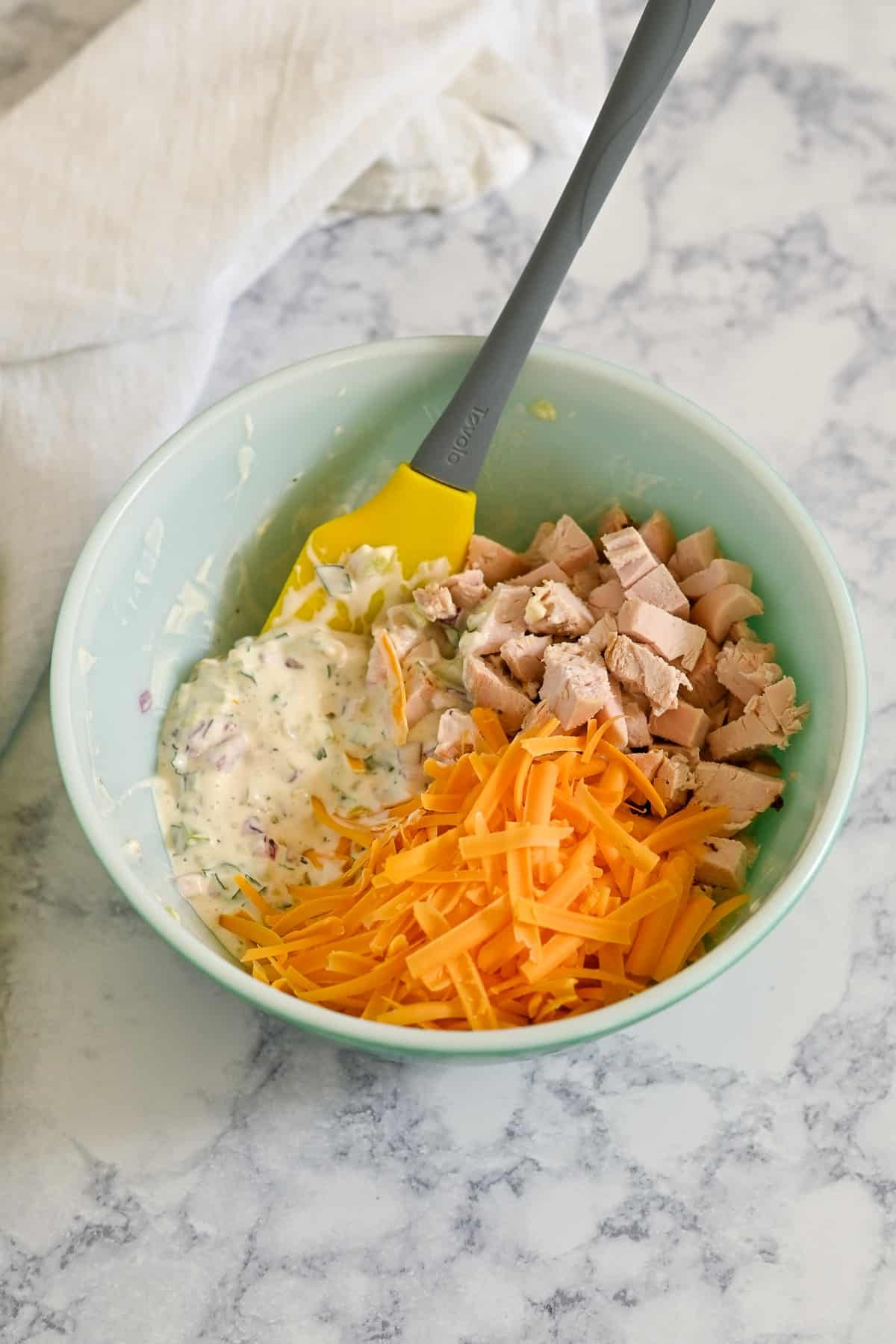 A bowl with chopped chicken, shredded cheddar, and creamy mixture being stirred with a spatula-perfect ingredients coming together for a delicious Rotisserie Chicken Macaroni Salad.
