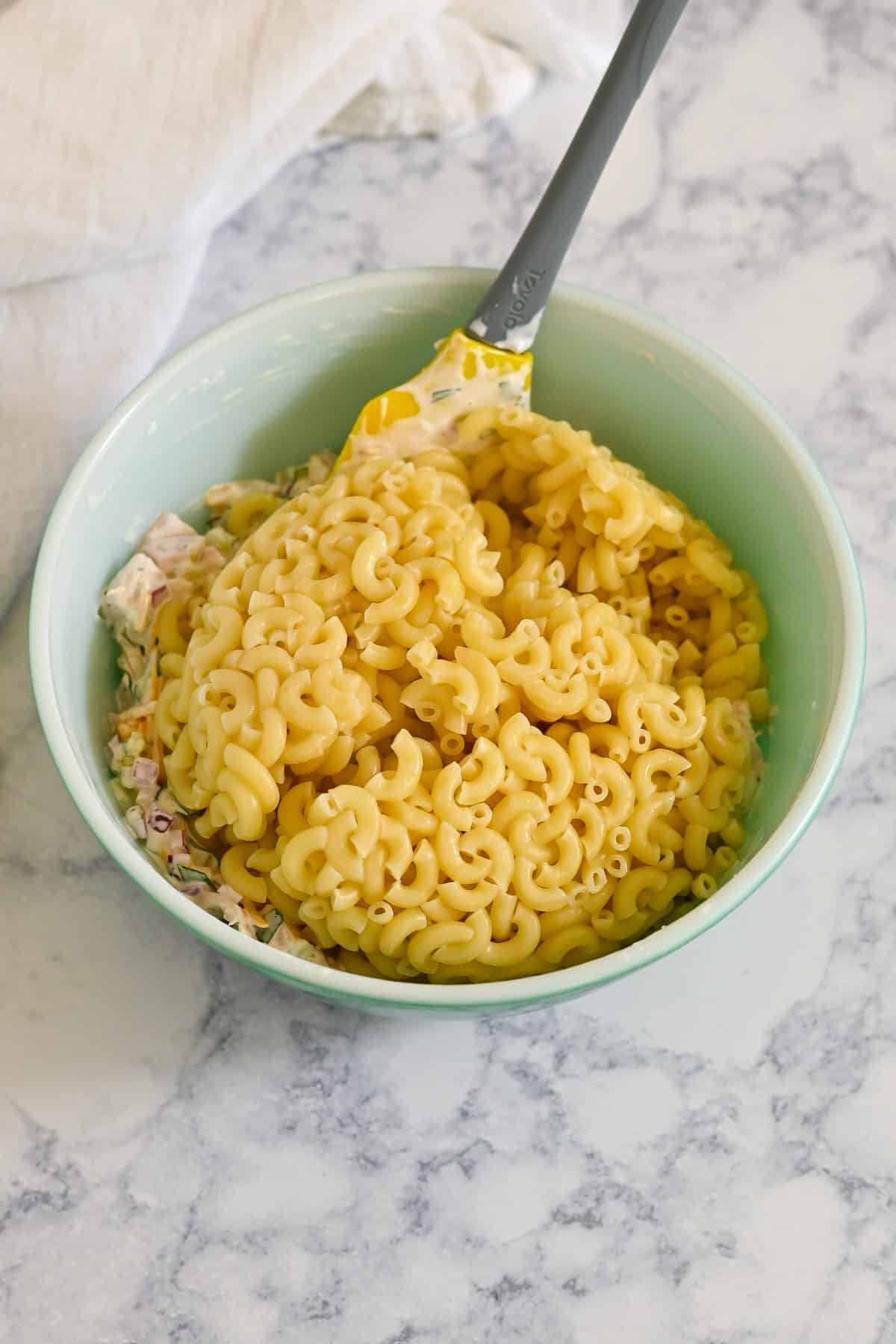 A bowl of cooked elbow macaroni on top of a creamy mixture, with a spatula in the bowl-perfect for preparing Rotisserie Chicken Macaroni Salad.