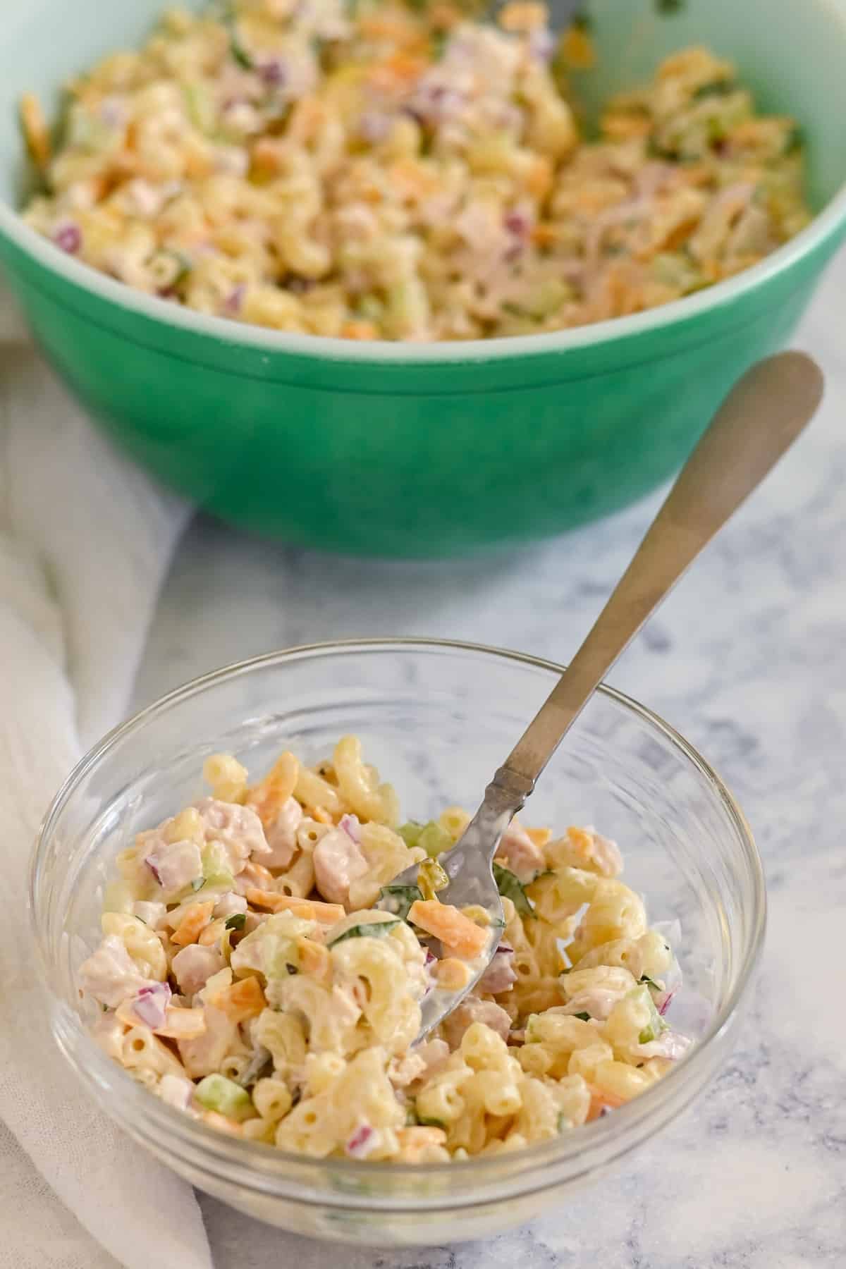 A glass bowl of chicken macaroni salad with vegetables and a fork sits in the foreground, while a large green bowl of salad is visible in the background.