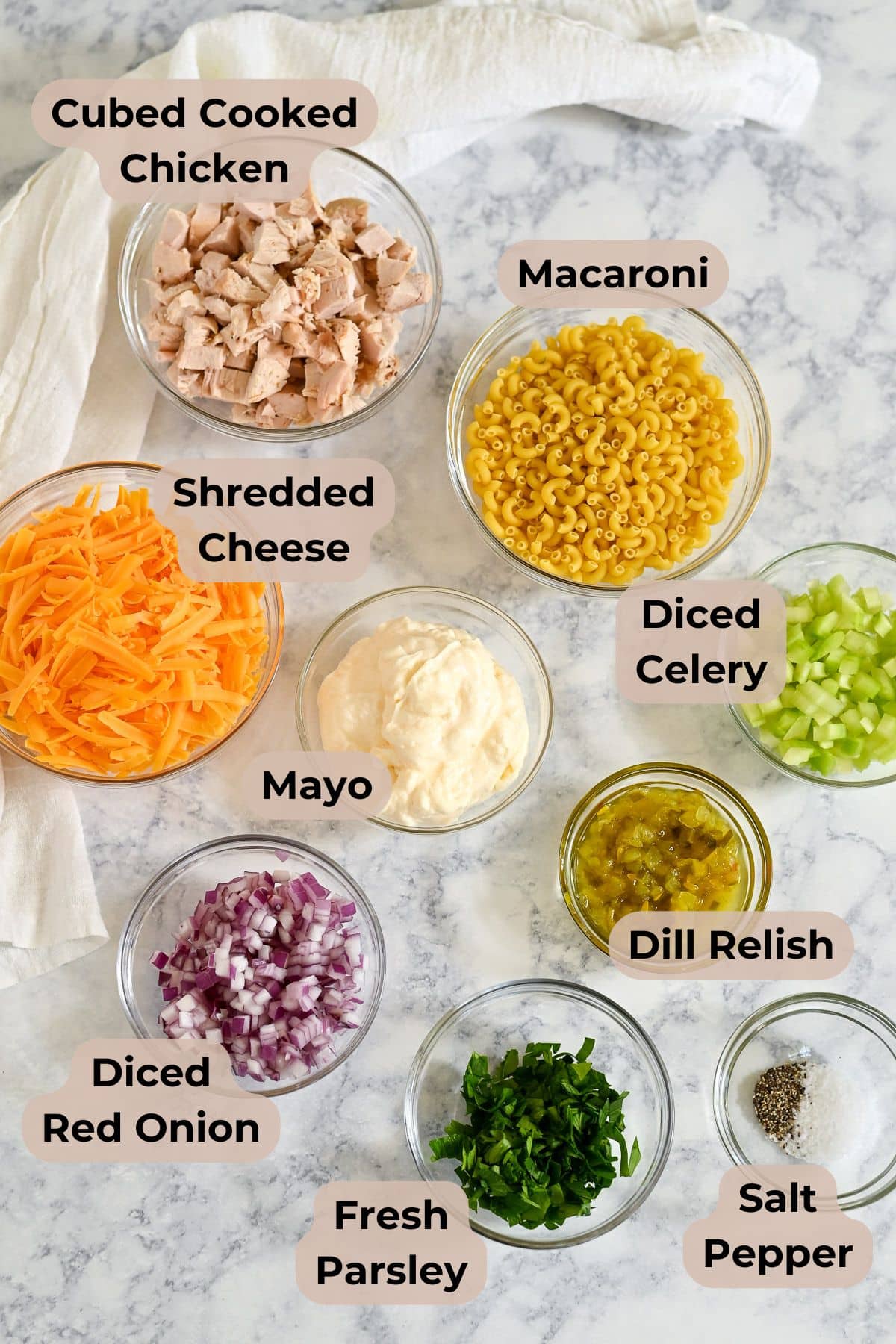 Bowls of cubed chicken, macaroni, cheese, mayo, celery, red onion, relish, parsley, salt, and pepper on a table-perfect for creating a delicious Rotisserie Chicken Macaroni Salad or your favorite chicken macaroni salad recipe.