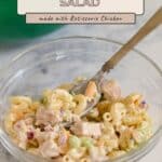 Clear bowl of chicken macaroni salad with tender rotisserie chicken, celery, and onion, with a fork inside, on a marble surface.