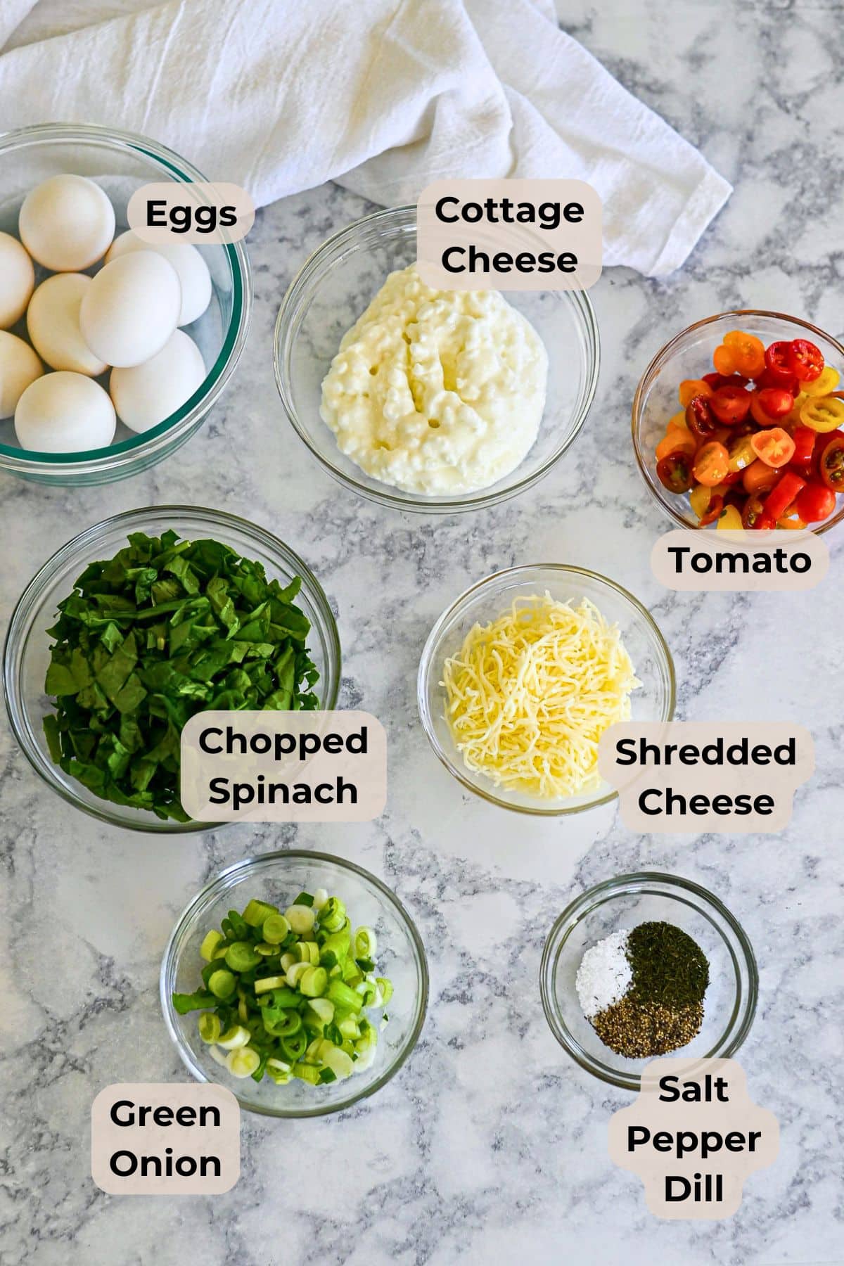 Seven bowls on a marble counter with ingredients for Spinach Egg Cups: eggs, cottage cheese, tomato, chopped spinach, shredded cheese, green onion, and spices.