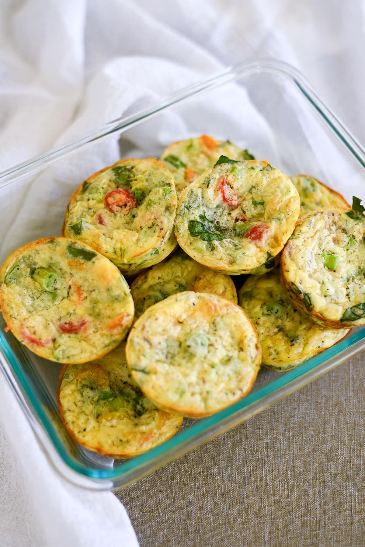 Mini Spinach Egg Cups in a glass container, topped with fresh herbs and vegetables, rest on a cloth napkin.
