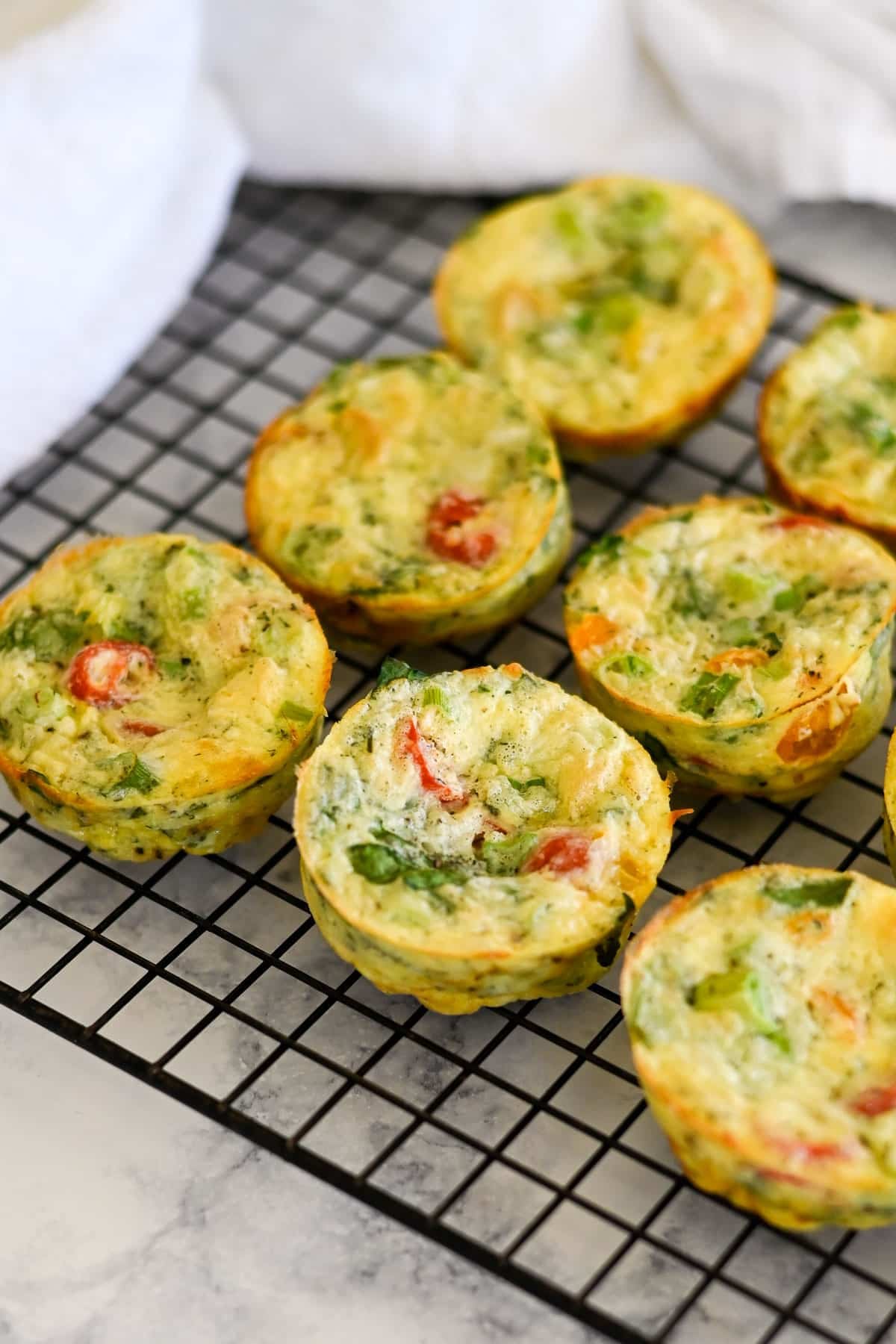 Mini Spinach Egg Cups and vegetable frittatas cooling on a black wire rack, with visible tomatoes and greens.