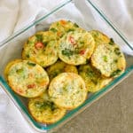 A glass container filled with mini vegetable egg muffins, including savory Spinach Egg Cups, sits on a beige surface next to a white cloth.