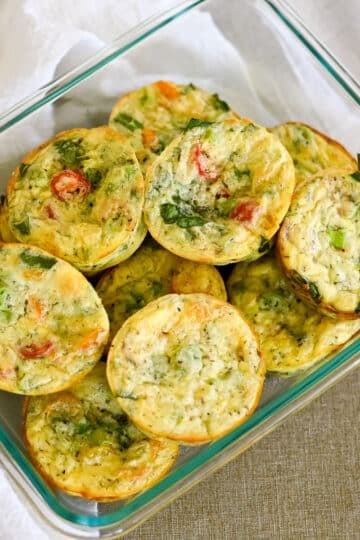 A glass container filled with mini vegetable egg muffins, including savory Spinach Egg Cups, sits on a beige surface next to a white cloth.