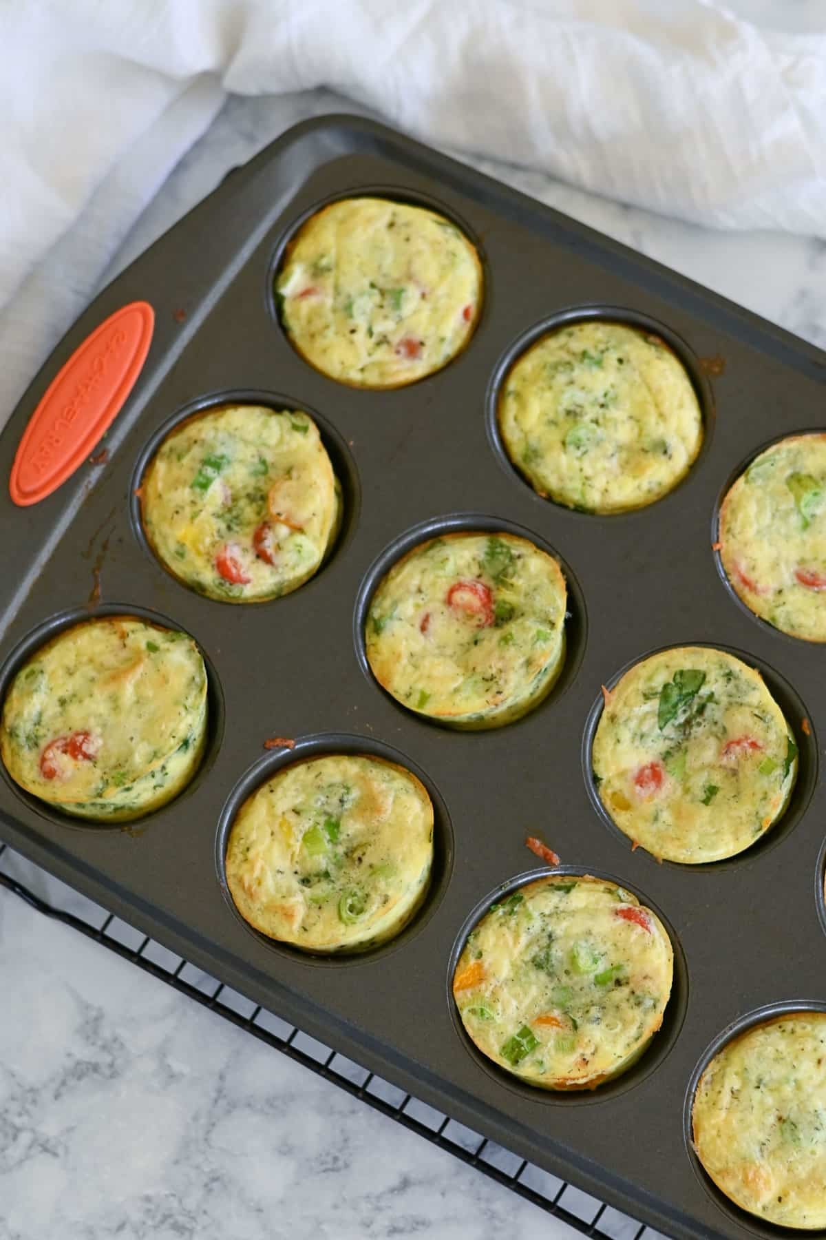 A muffin tin filled with freshly baked Spinach Egg Cups with vegetables sits on a marble countertop.