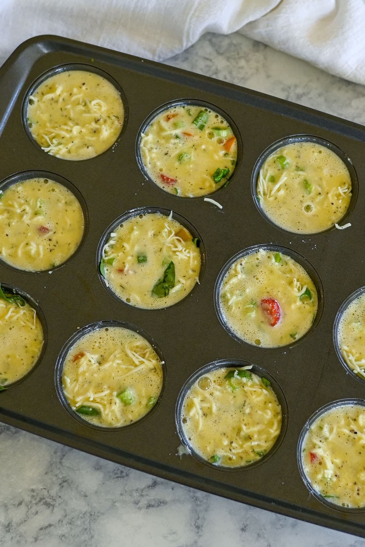 Muffin pan filled with uncooked egg mixture, veggies, cheese, and fresh spinach, ready to be baked into delicious Spinach Egg Cups.