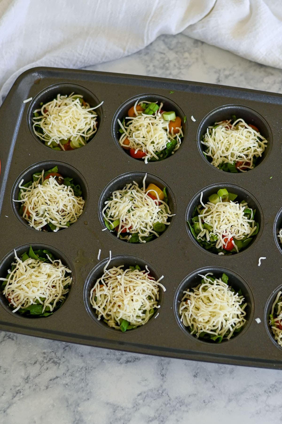 A muffin tin filled with chopped vegetables and shredded cheese, ready to be baked into delicious Spinach Egg Cups.